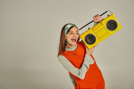 mujer llena de alegría en naranja vestido de estilo retro sosteniendo boombox amarillo y bailando en gris, felicidad
