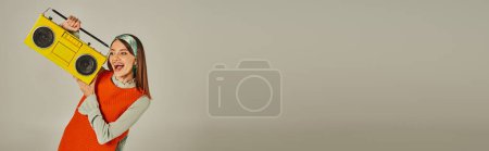 Photo for Cheerful woman in stylish orange dress holding yellow vintage boombox and dancing on grey, banner - Royalty Free Image