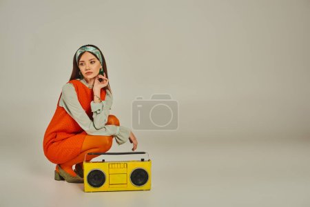 thoughtful woman in orange dress listening music on yellow boombox on grey, retro-inspired lifestyle