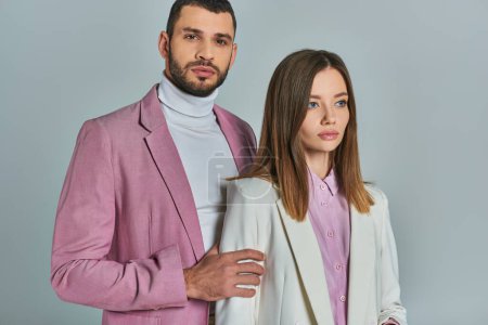homme confiant en blazer lilas regardant la caméra près de la femme en costume blanc sur gris, mode d'affaires