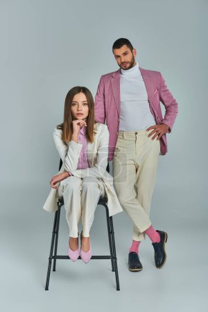 confident man in lilac blazer with hand on hip near woman in white suit sitting on chair on grey
