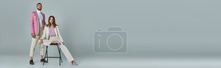Téléchargez les photos : Jeune femme élégante en costume blanc posant sur chaise près de l'homme confiant en blazer lilas sur gris, bannière - en image libre de droit