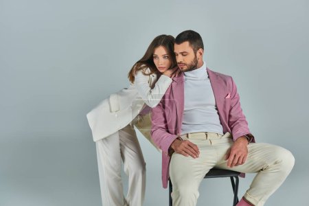 young woman in white suit leaning on confident man in lilac blazer sitting on chair on grey backdrop