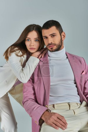 Frau im weißen Anzug stützt sich auf einen selbstbewussten Mann im fliederfarbenen Blazer, der sitzt und in die Kamera auf grau blickt