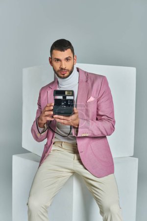 handsome and stylish man in lilac blazer holding vintage camera near white cubes on grey backdrop