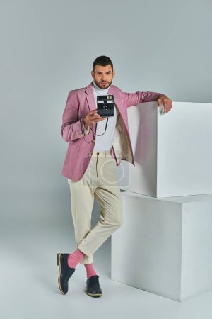 stylish man in lilac blazer and white pants taking photo on vintage camera near cubes on grey