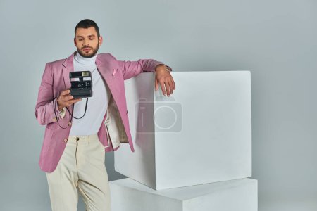 joven en ropa formal elegante posando cerca de cubos blancos con cámara vintage en gris, moda moderna