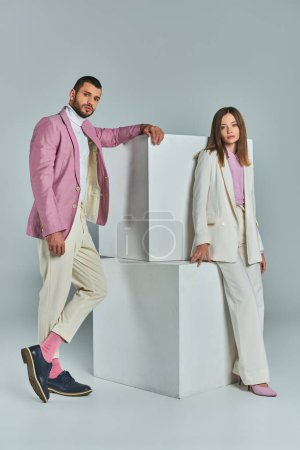 Téléchargez les photos : Jeune couple confiant en costumes à la mode posant près de cubes blancs sur gris, mode minimaliste - en image libre de droit