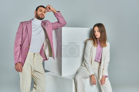 Trendiger Mann in fliederfarbenem Blazer blickt in die Kamera neben weißen Würfeln und stilvolle Frau sitzt auf grau