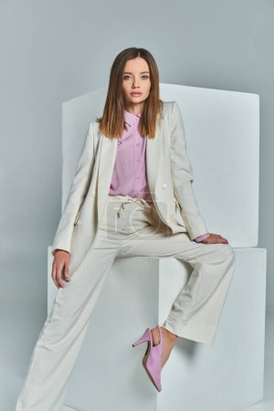 attraktive Frau im eleganten Anzug posiert neben weißen Würfeln auf grauer, minimalistischer Businessmode