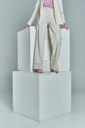 vista recortada de mujer joven en elegante atuendo formal de pie sobre cubos blancos sobre fondo gris