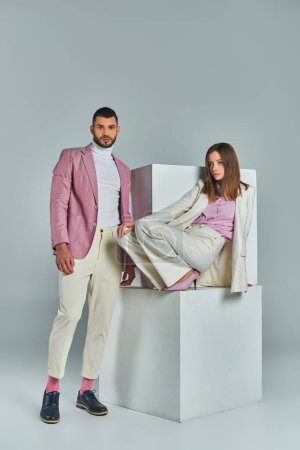 Photo for Fashionable man in lilac blazer looking at camera near woman in white suit posing on cubes on grey - Royalty Free Image