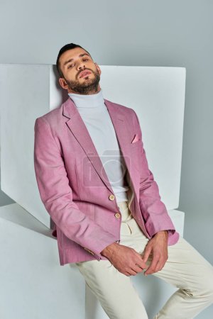 hombre carismático en blazer lila de moda sentado en cubo blanco y mirando a la cámara en gris