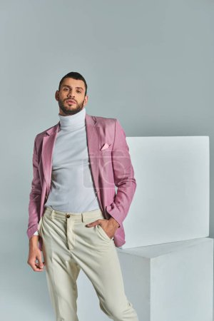 hombre seguro en chaqueta lila posando con la mano en el bolsillo cerca de cubos blancos en gris, moda de negocios