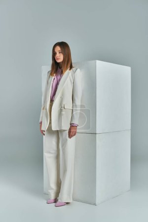 trendy business attire, modern woman in white elegant suit standing near cubes on grey, full length