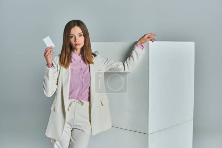 young successful woman in stylish formal attire holding blank business card near white cubes on grey