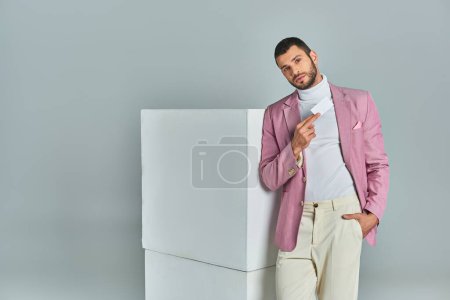 hombre de moda en blazer lila posando con la mano en el bolsillo y la tarjeta de visita en blanco cerca de cubos en gris