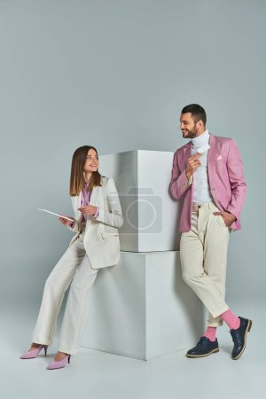 Stilvolles Paar mit Visitenkarte und digitalem Tablet lächelt einander in der Nähe von Würfeln auf grau an