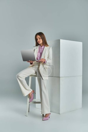 joven mujer de negocios en traje elegante usando el ordenador portátil en el taburete cerca de cubos blancos en gris, longitud completa