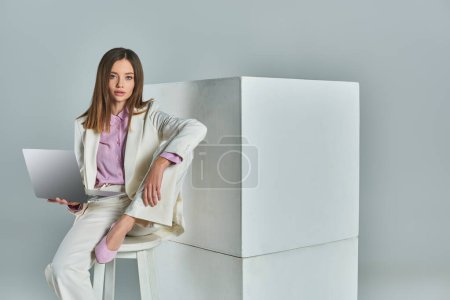 Elegante Frau im weißen Anzug sitzt auf einem Hocker mit Laptop und blickt in die Kamera in der Nähe von Würfeln auf grau