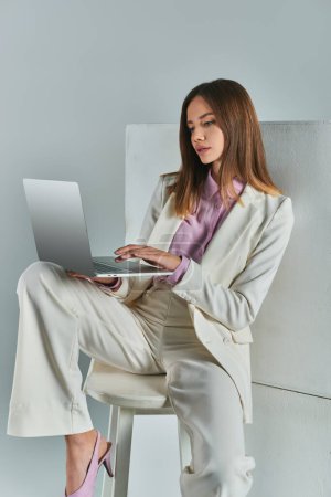 young woman in elegant suit networking on laptop while sitting on stool near white cubes on grey