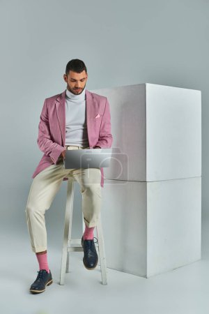 junger Geschäftsmann in fliederfarbenem Blazer sitzt auf einem Hocker und arbeitet am Laptop neben weißen Würfeln auf grau