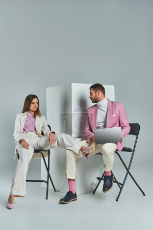 Stylischer Mann mit Laptop und junge Frau im weißen Anzug sitzen auf Stühlen neben Würfeln vor grauem Hintergrund