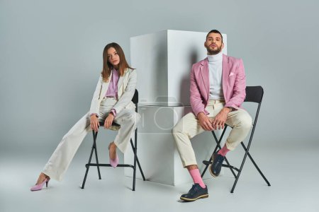 Photo for Confident couple in stylish formal wear sitting on chairs near cubes on grey, minimalistic fashion - Royalty Free Image