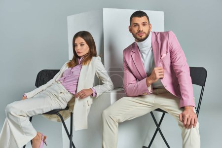 junges Paar in angesagter formaler Kleidung sitzt auf Stühlen neben Würfeln auf grauer, moderner Businessmode