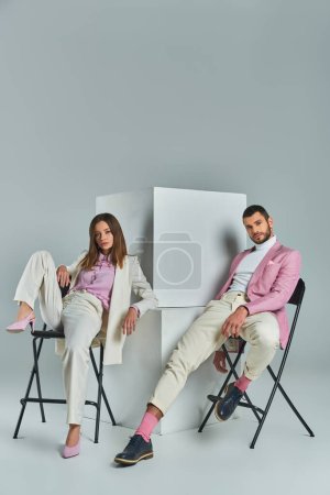 junges Paar in stylischer Businesskleidung auf Stühlen neben Würfeln auf grauer, minimalistischer Mode sitzend