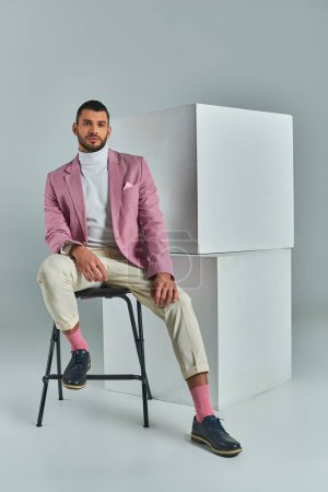 hombre elegante en traje de negocios sentado en la silla y mirando a la cámara cerca de cubos blancos en gris