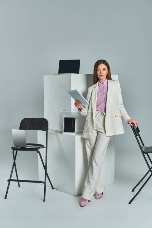 junge Frau im weißen Anzug mit Laptop in der Nähe von Stühlen und digitalen Geräten auf weißen Würfeln auf grau
