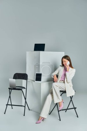 mujer sonriente sentada en la silla y hablando en el teléfono inteligente cerca de dispositivos en cubos blancos en gris