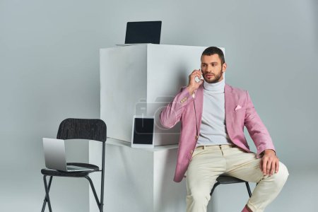 confident stylish man talking on smartphone while sitting near devices on white cubes on grey