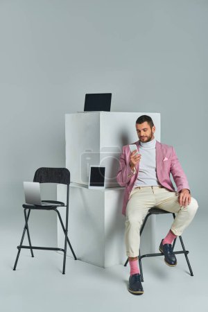 homme élégant en blazer lilas assis sur une chaise avec smartphone près des appareils sur cubes blancs sur gris