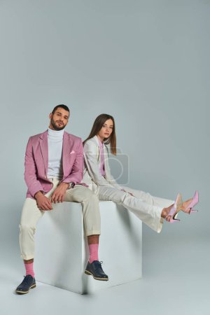 junges Businesspaar in eleganter formaler Kleidung sitzt auf einem weißen Würfel und blickt in die Kamera auf grau