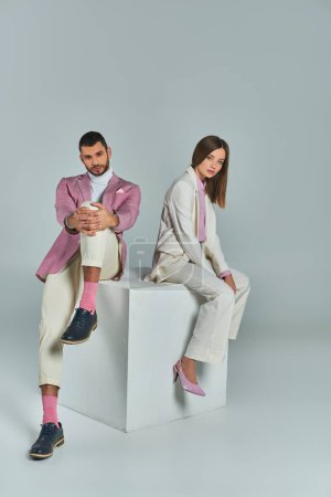 trendiges Businesspaar in eleganter, formeller Kleidung sitzt auf einem White Cube und blickt in die Kamera auf grau