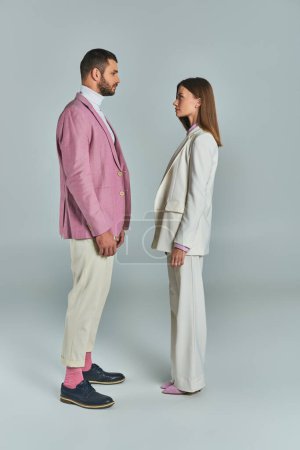Téléchargez les photos : Longueur totale de jeune couple en costumes élégants se regardant sur gris, mode d'affaires moderne - en image libre de droit