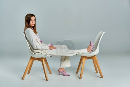 mujer joven y elegante en traje blanco posando en sillones en gris, moda de negocios minimalista