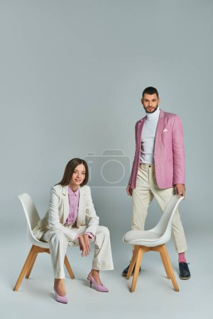 glückliche Frau im weißen Anzug sitzt im Sessel neben einem Mann im lila Blazer und blickt in die Kamera auf grau