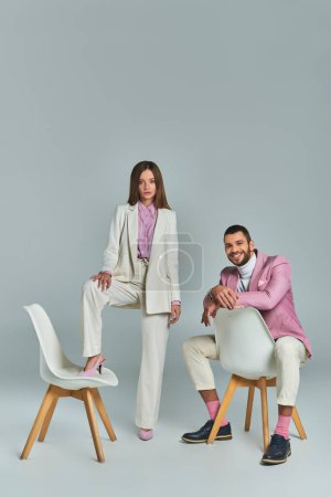 joyful man in lilac blazer sitting and looking at camera near woman posing with armchair on grey
