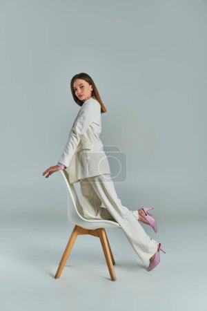mujer moderna en traje elegante blanco mirando a la cámara y posando con sillón en gris, longitud completa