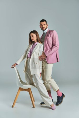 junges elegantes Paar in pastellfarbener formaler Kleidung posiert mit Sessel auf grauem, modernem Businessstil