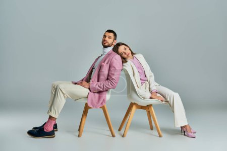 Foto de Joven pareja en elegante formal desgaste sentado espalda con espalda en sillones y mirando a la cámara en gris - Imagen libre de derechos