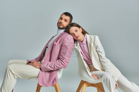 elegant couple in formal wear sitting in armchairs and looking at camera on grey, modern fashion
