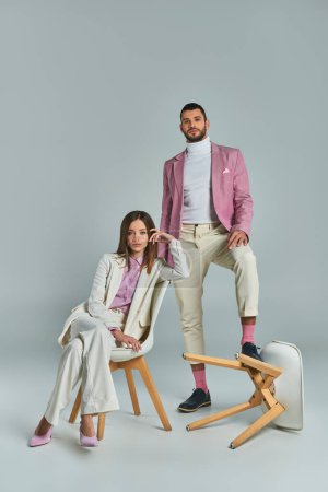 Modisches Paar in eleganter, formaler Kleidung posiert mit Sesseln auf grauer, moderner Businessmode