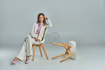 pleine longueur de femme moderne en costume blanc assis sur un fauteuil et regardant la caméra sur le gris