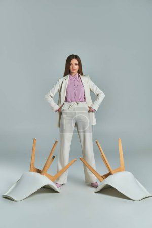 Selbstbewusste Geschäftsfrau im weißen Anzug steht mit den Händen auf den Hüften neben umgestürzten Sesseln auf grau