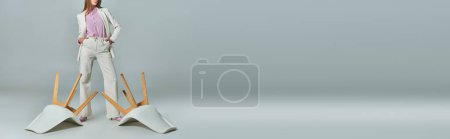 Photo for Cropped view of woman in white suit with hands in pockets near overturned armchairs on grey, banner - Royalty Free Image