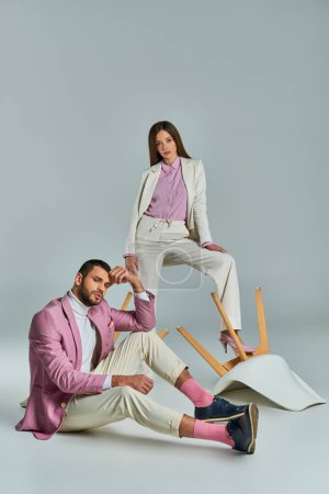 Modisches Paar in eleganter, formaler Kleidung posiert mit umgestürzten Sesseln auf grauer, moderner Mode